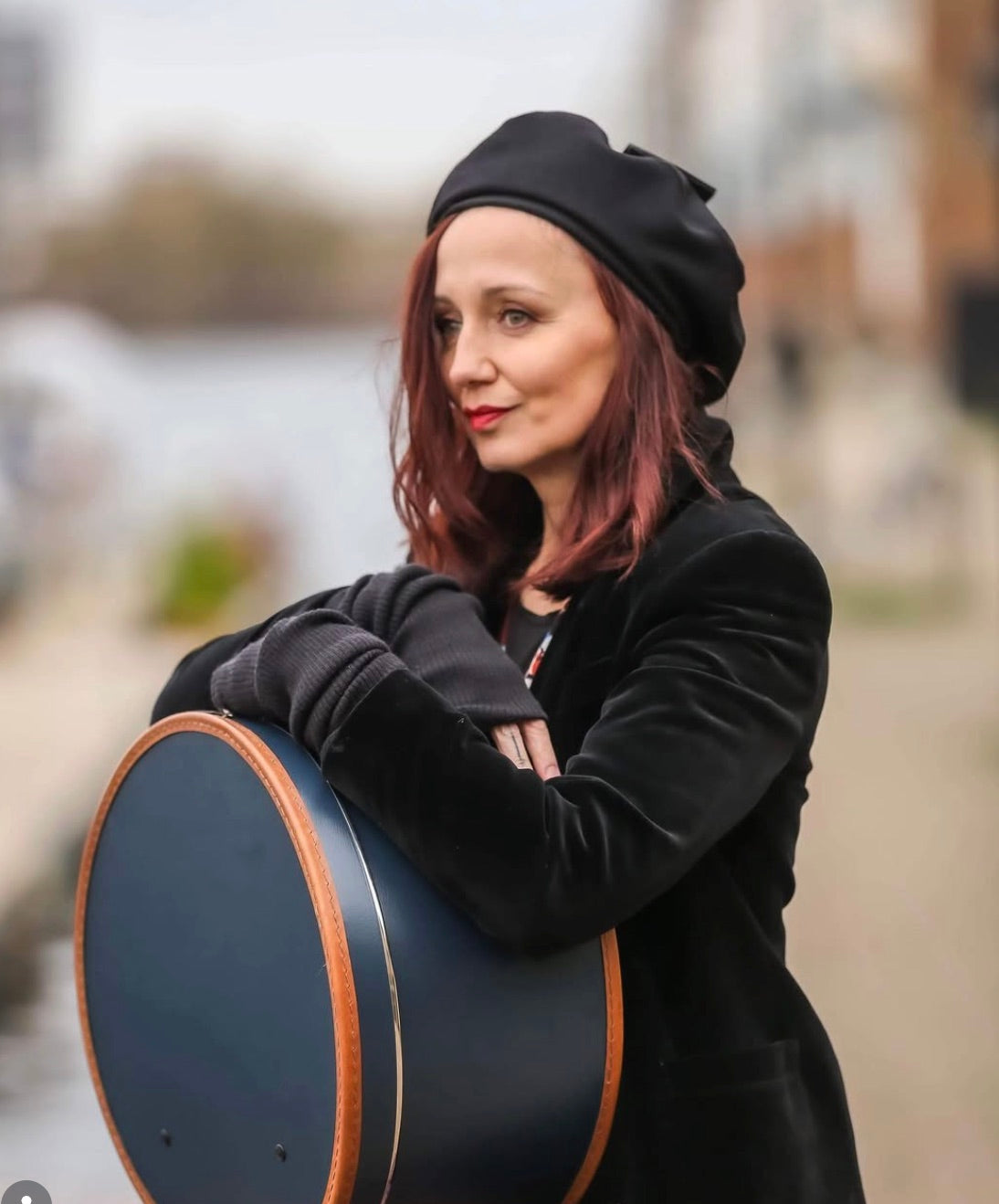 Woman in a black beret and velvet coat carrying a navy hat box beside a river