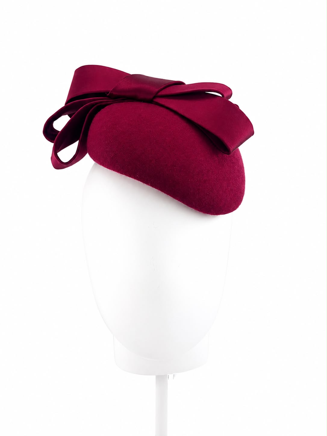 Claret wool crêpe cocktail hat with a sculptural silk bow, worn to one side on a head block, Maria Monica London