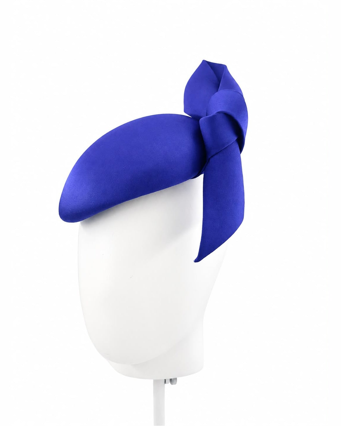 Cobalt blue satin cocktail hat with a sculptural knotted bow, shown on a white display head, Maria Monica London
