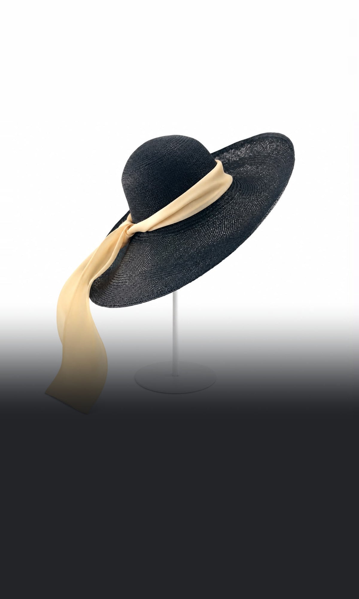 Black wide brim straw hat with a cream silk sash trailing from the brim, on a stand, Maria Monica London