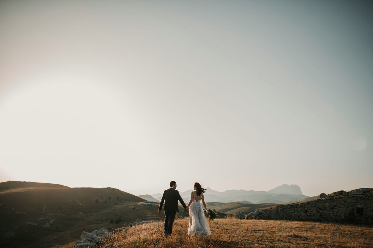 A couple in wedding clothes walking hand in hand across a dry hillside at sunset, mountains behind