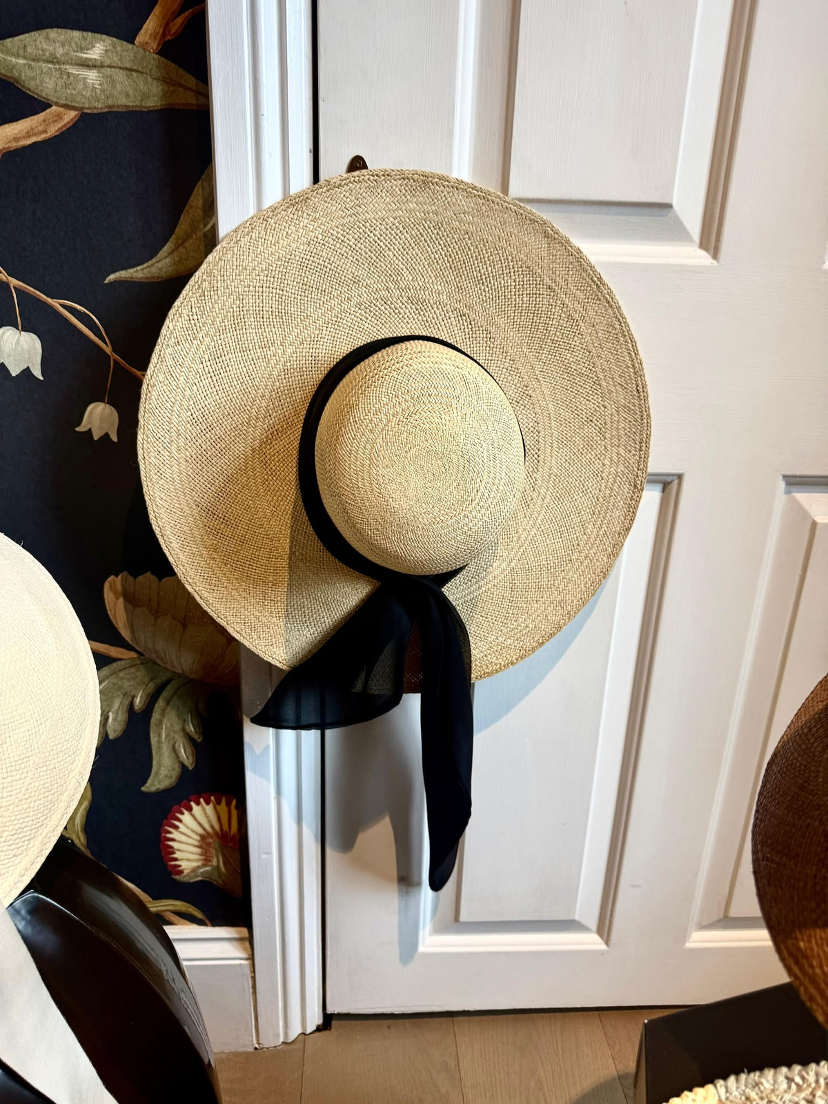 Natural straw wide brim hat with a black chiffon scarf tie, hanging on a white panelled door