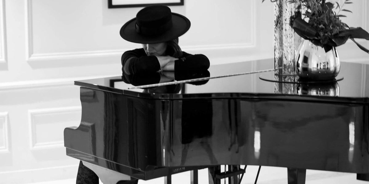 Black and white photograph of a woman in a wide brim black hat leaning on a grand piano