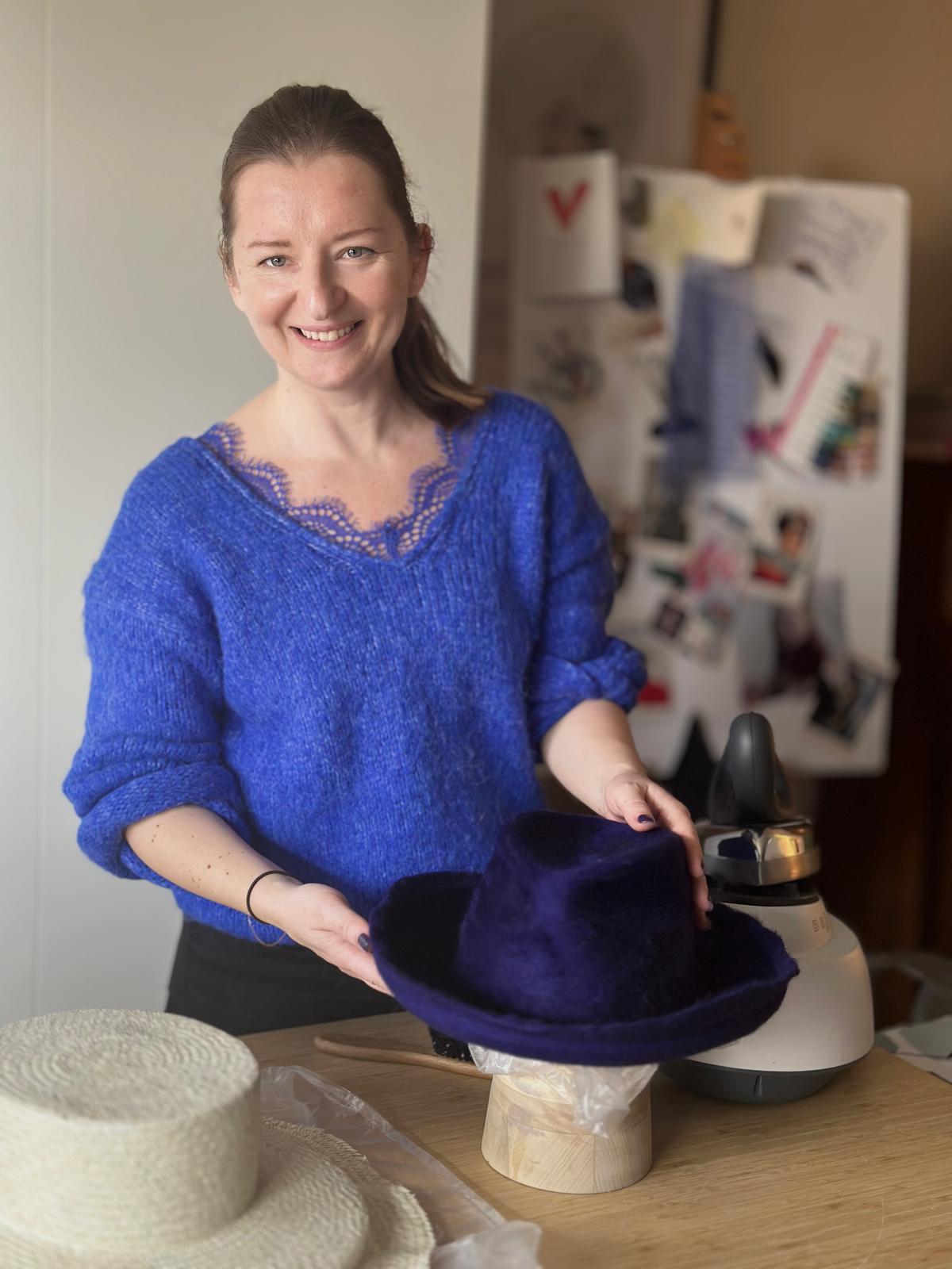 A woman at a workbench holding a purple felt hat on a wooden block beside a steamer