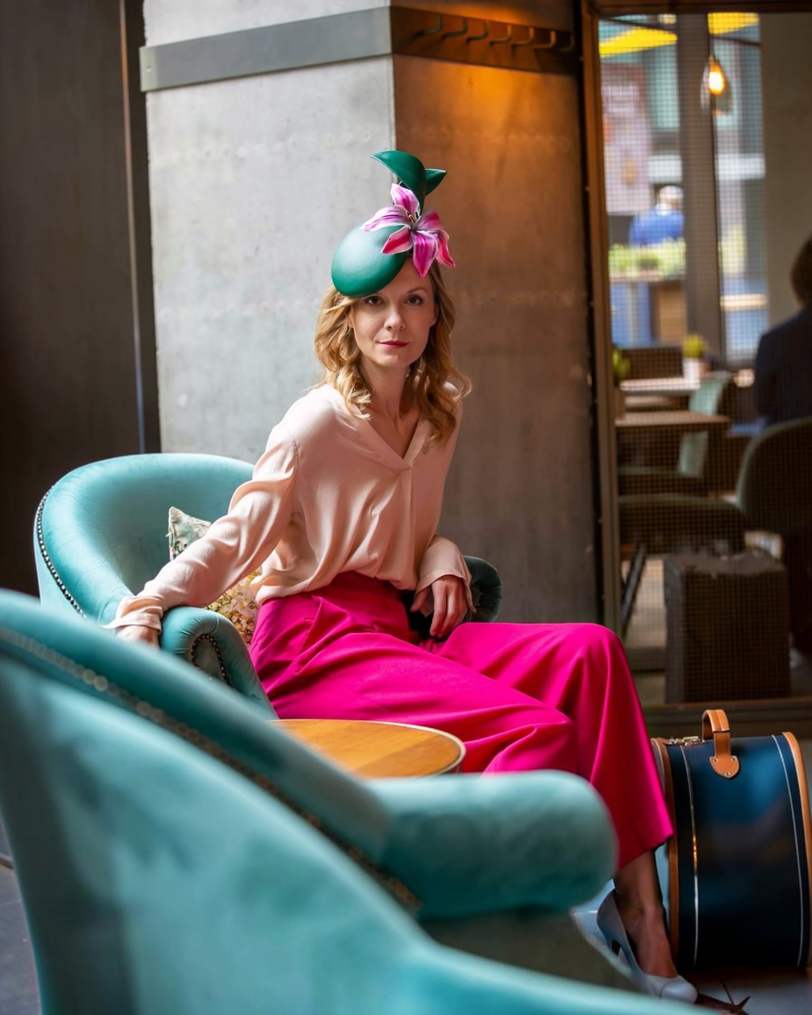 Woman seated in a hotel lounge wearing the emerald cocktail hat with a pink lily, Maria Monica London