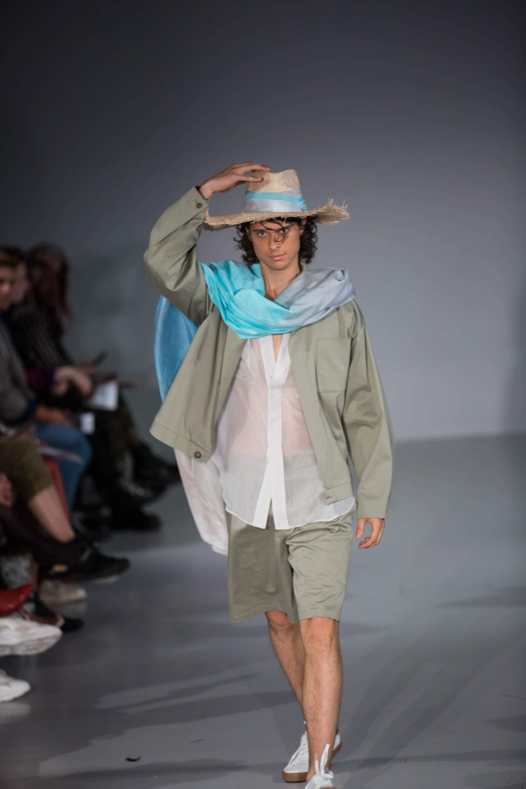 Model walking a runway in a frayed brim straw hat with a pale blue band and a sage jacket