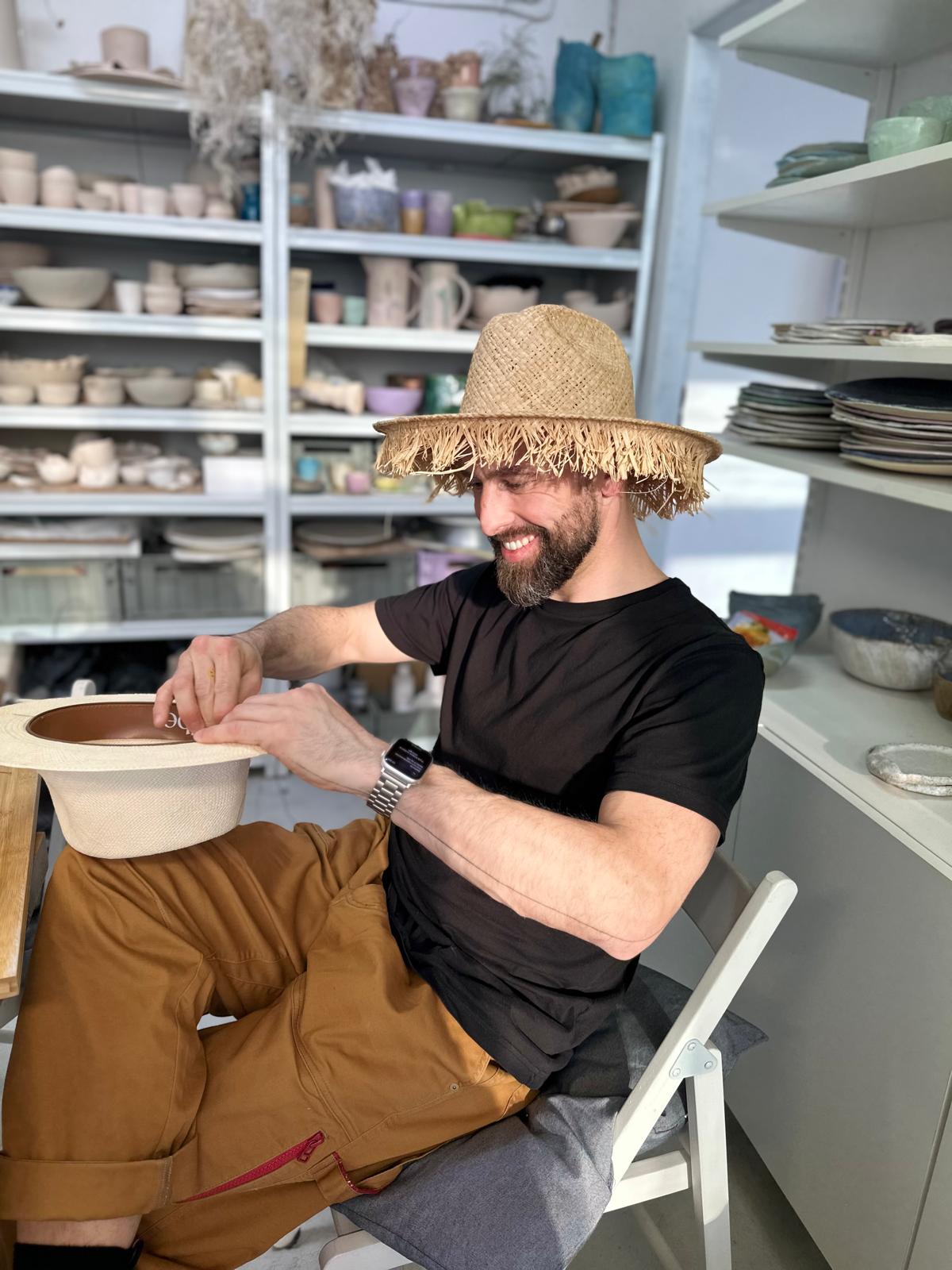 A person in a frayed raffia hat shaping the brim of a cream straw hat, shelves of pottery behind