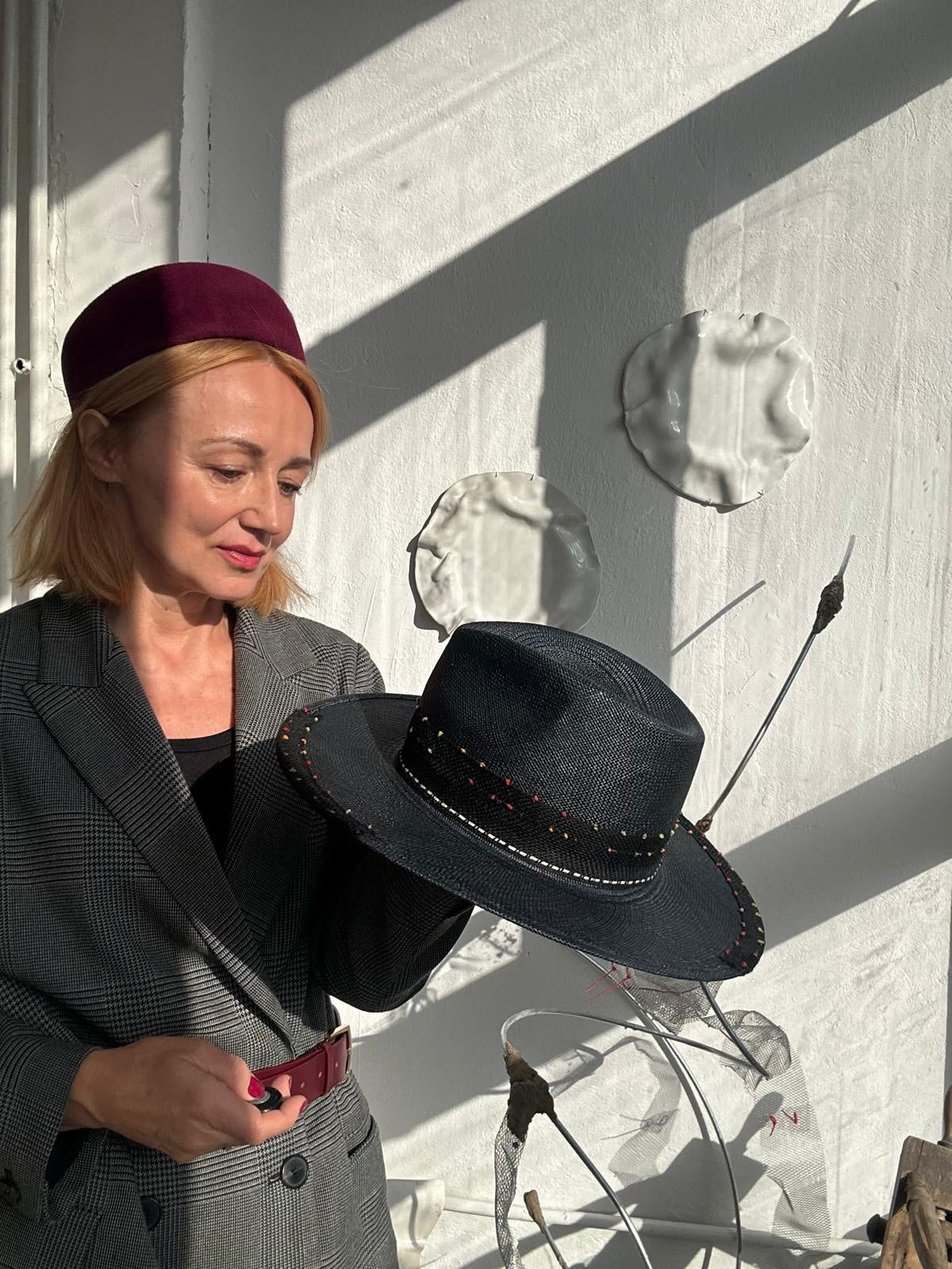 Woman in a burgundy pillbox hat holding a black straw fedora with a stitched band in a sunlit studio