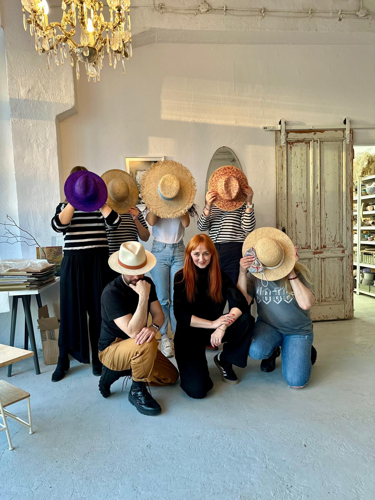 Millinery workshop group in a white studio holding straw and felt hats in front of their faces