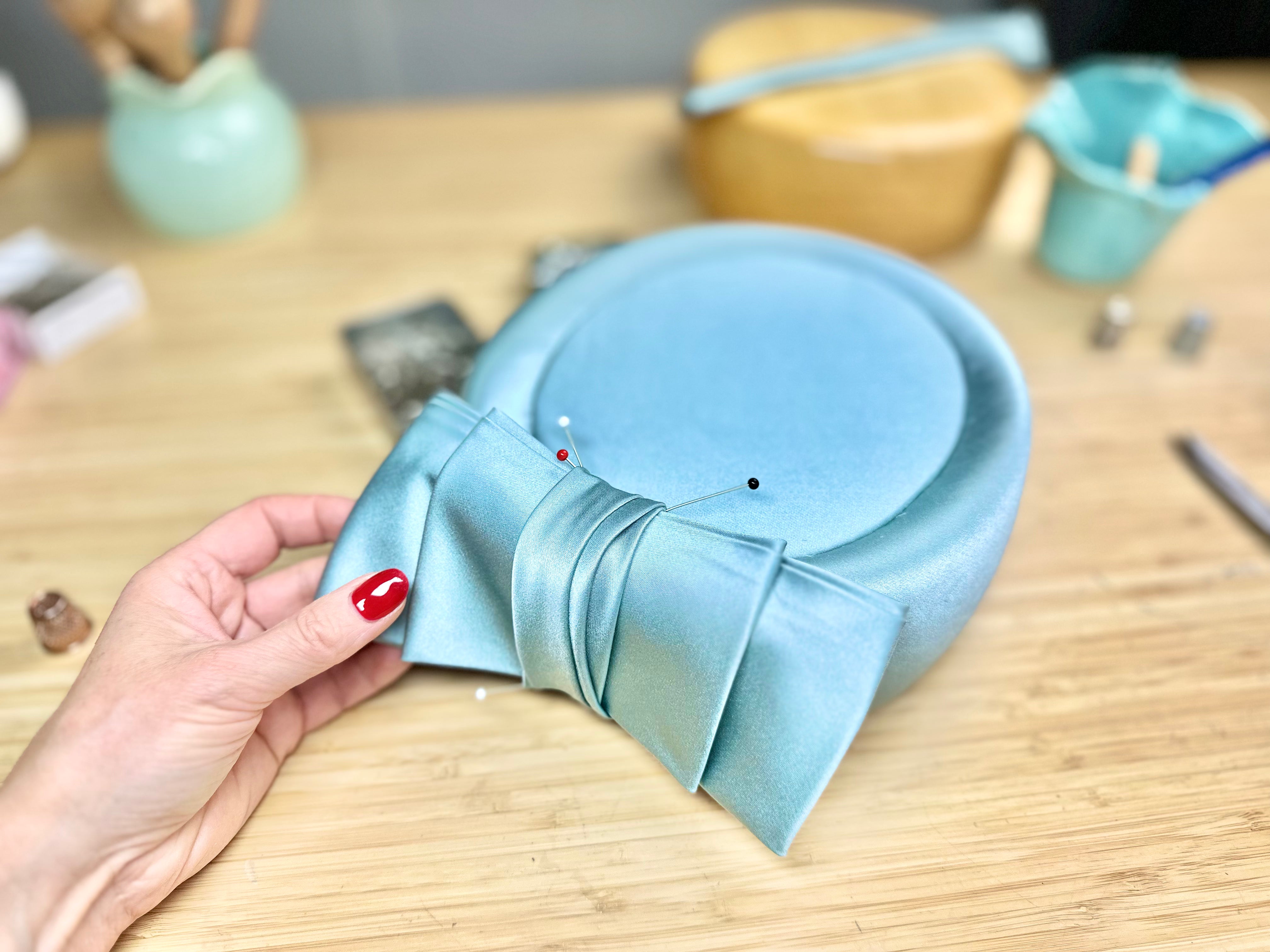 A hand holding a pale blue pillbox hat in progress with a satin bow pinned on a workbench