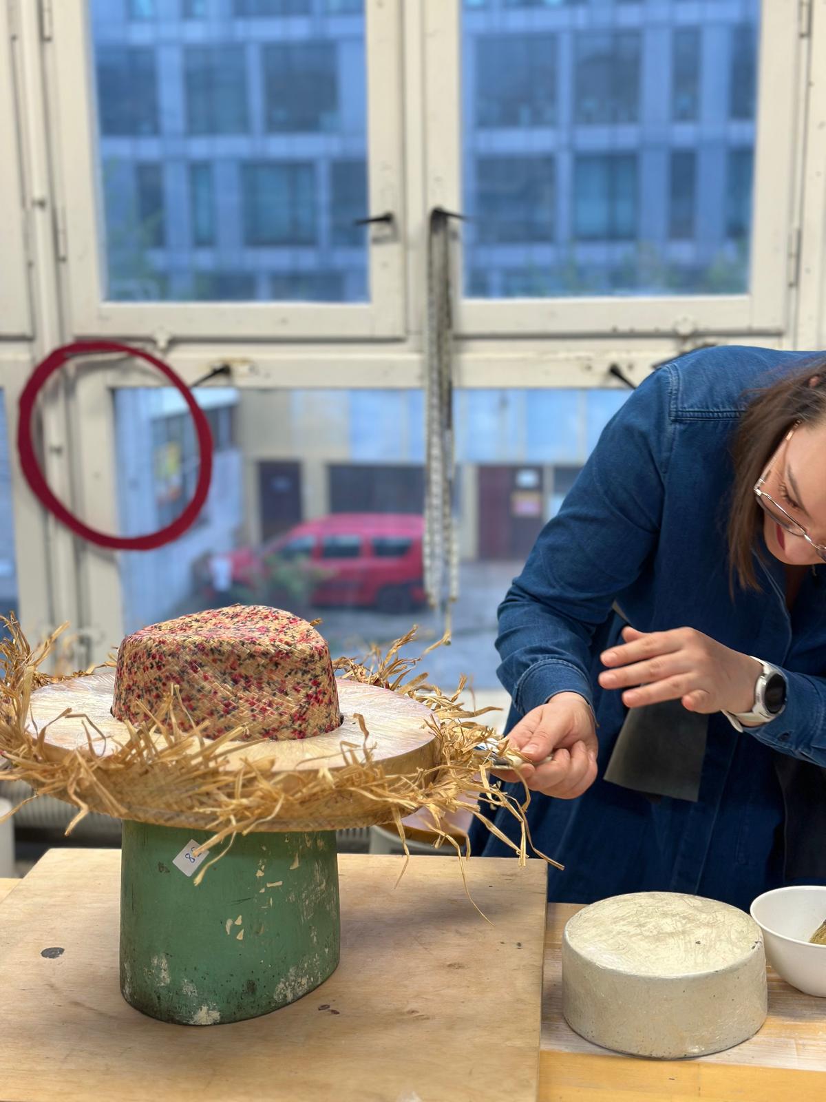 Milliner working loose straw at the brim of a red flecked hat on a wooden block