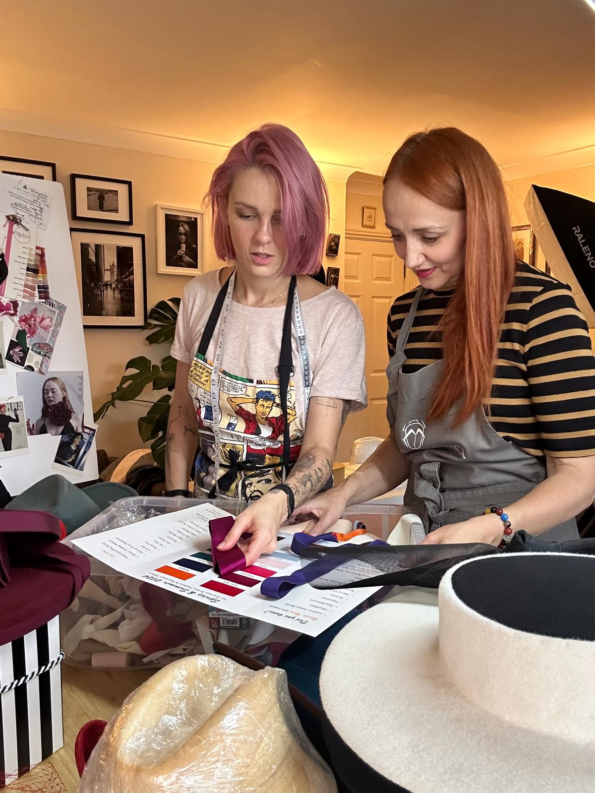 Two milliners in aprons looking at a printed colour card with ribbon and felt swatches in a workroom